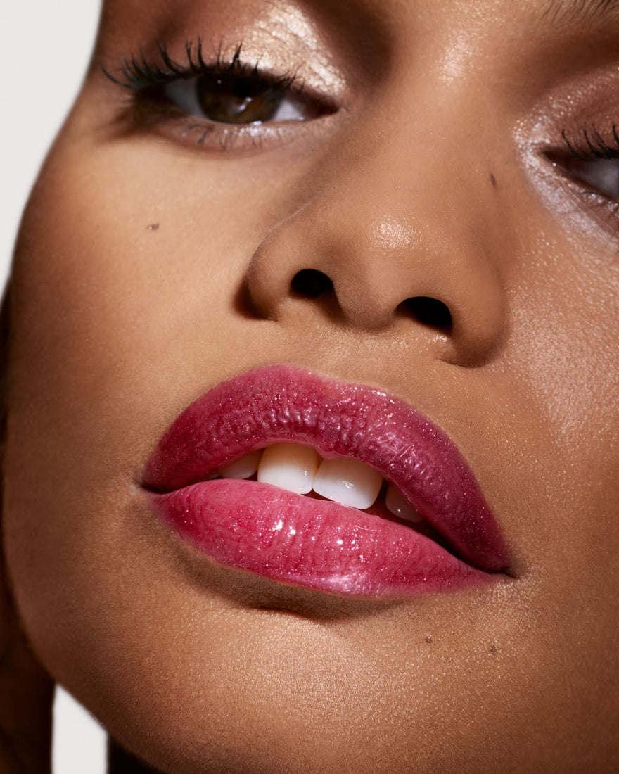 Close-up of a medium deep-toned model's face wearing Gloss Bomb Universal Lip Luminizer in the shade Fuchsia Flex.