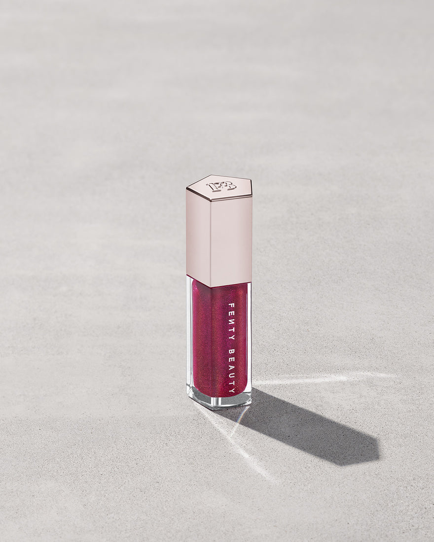 A closed tube of Gloss Bomb Universal Lip Luminizer in the shade Fuschia Flex on a grey background.